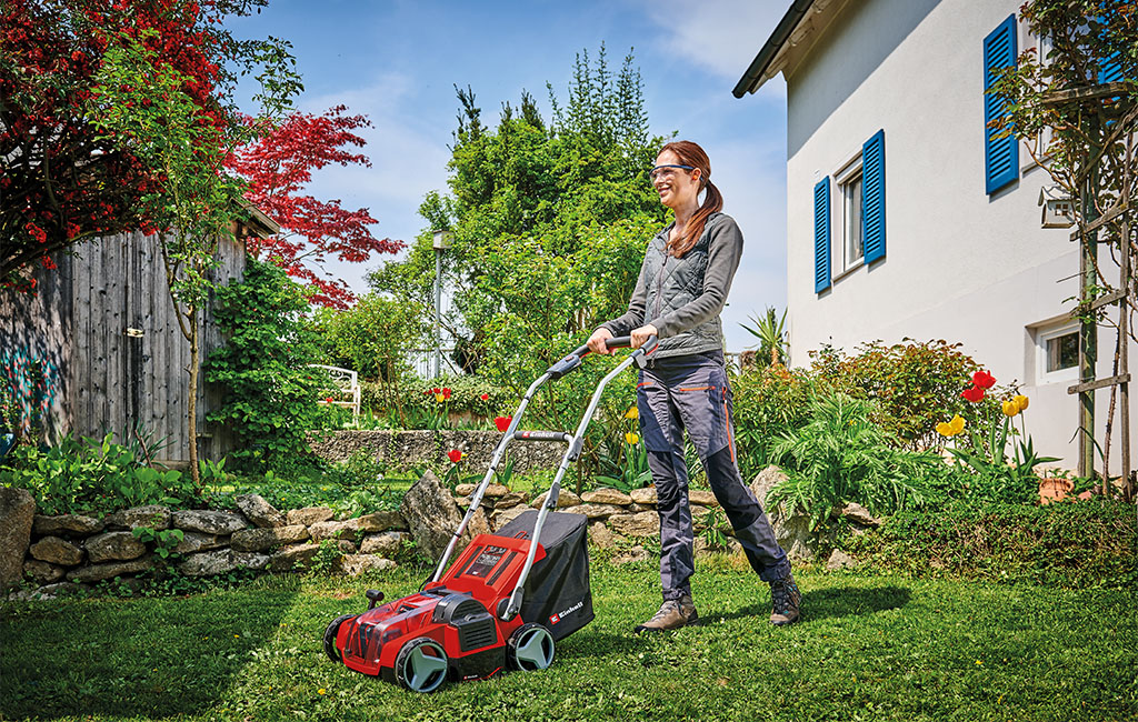 A woman aerating the lawn with a cordless scarifier aerator in the garden in front of a house.