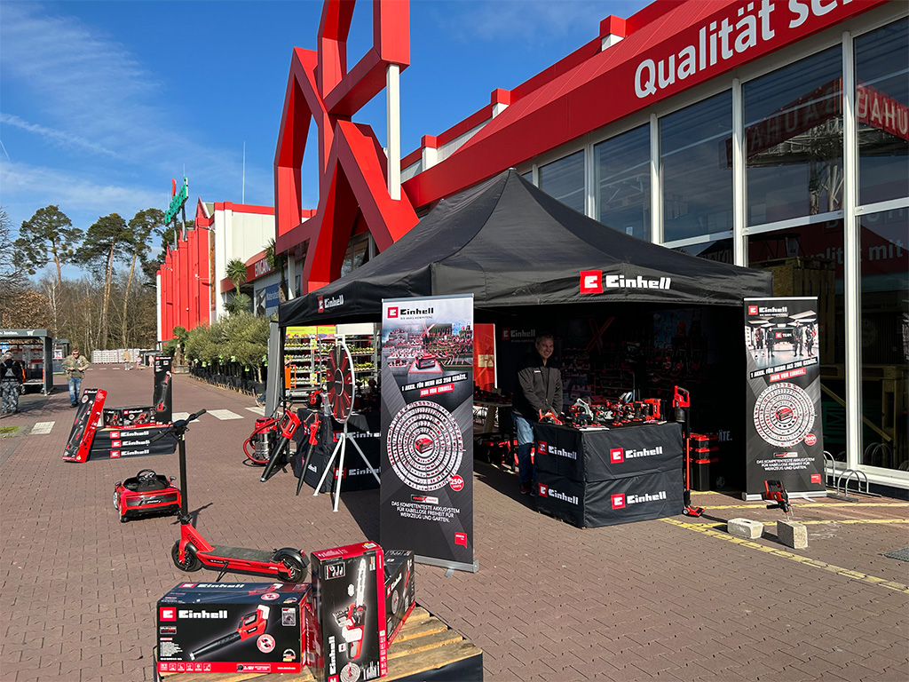 Einhell booth outside a hardware store, showcasing tools and products with banners and a spinning wheel for interaction.